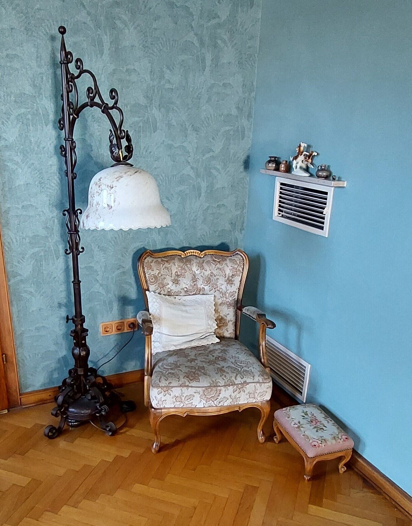 A room with a vintage-style chair, a tall floor lamp, a small bench, and a wall-mounted shelf with decorative items.