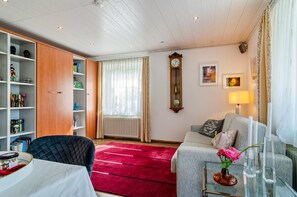 A living room with a red carpet, a sofa, a bookshelf, a radiator, a wall clock, and a window with curtains. - Apartment 'Fewo Wegen' with Mountain View, Private Terrace & Wi-Fi (Salem)