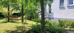 Ein Garten mit einer Holzbank, einem Baum und einem Gebäude mit Fenster und Balkon.