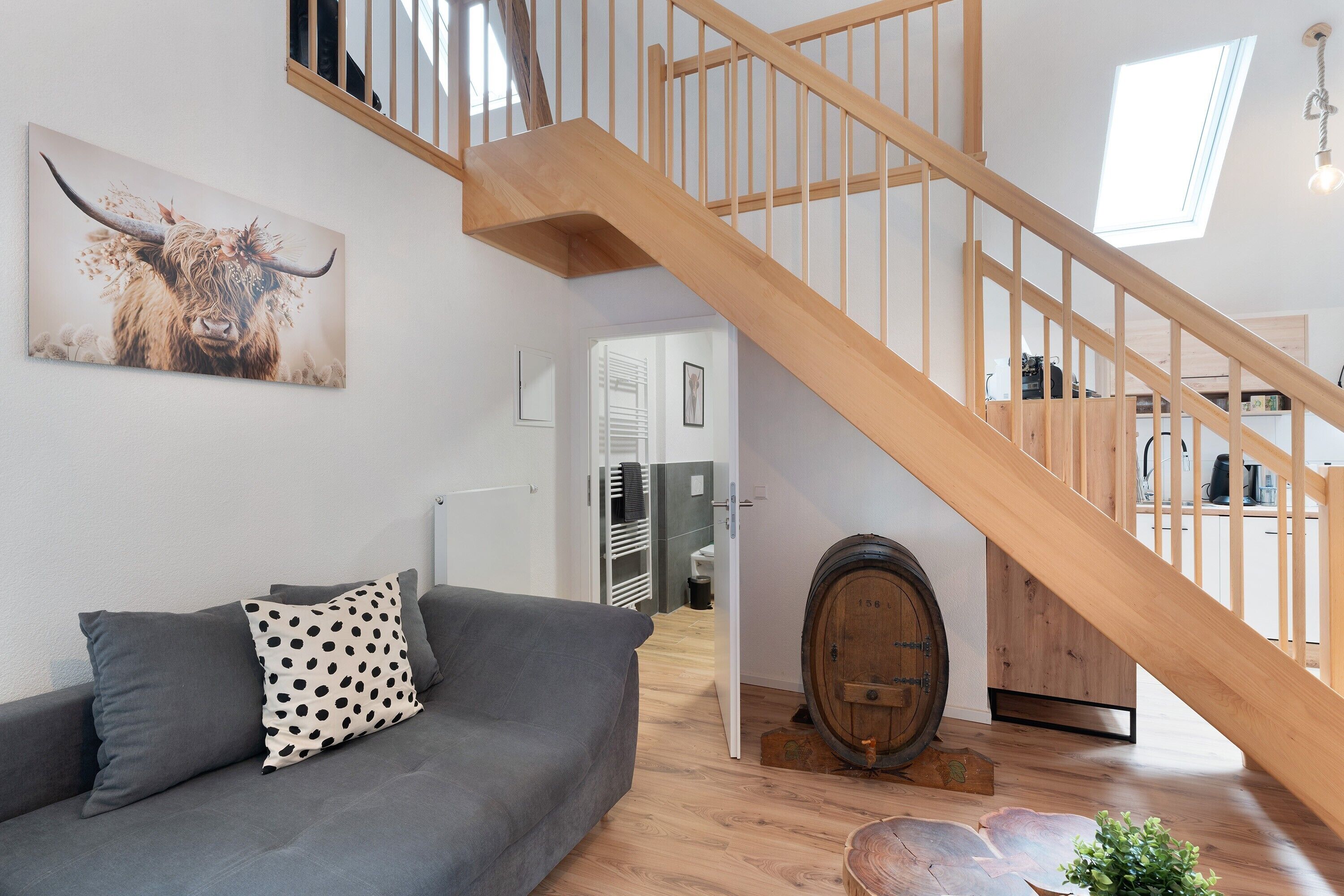 A staircase with wooden railings, a grey sofa with a polka dot pillow, a wooden barrel, and a bull painting on the wall.
