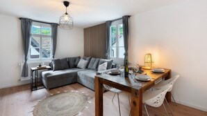 A modern living room with a grey sectional sofa, a wooden dining table with chairs, a round woven rug, and a pendant light. - Apartment 'Appartement Waldblick' with Mountain View, Shared Garden and Wi-Fi (Bühlertal)