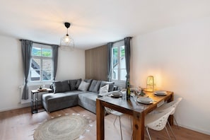 A modern living room with a grey sectional sofa, a wooden dining table with chairs, a round woven rug, and a pendant light. - Apartment 'Appartement Waldblick' with Mountain View, Shared Garden and Wi-Fi (Bühlertal)
