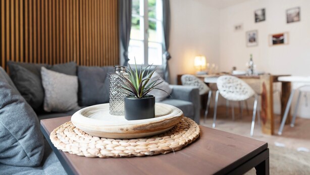 A living room with a sofa, a coffee table with a woven placemat, a potted plant, and a dining area in the background. - Apartment 'Appartement Waldblick' with Mountain View, Shared Garden and Wi-Fi (Bühlertal)