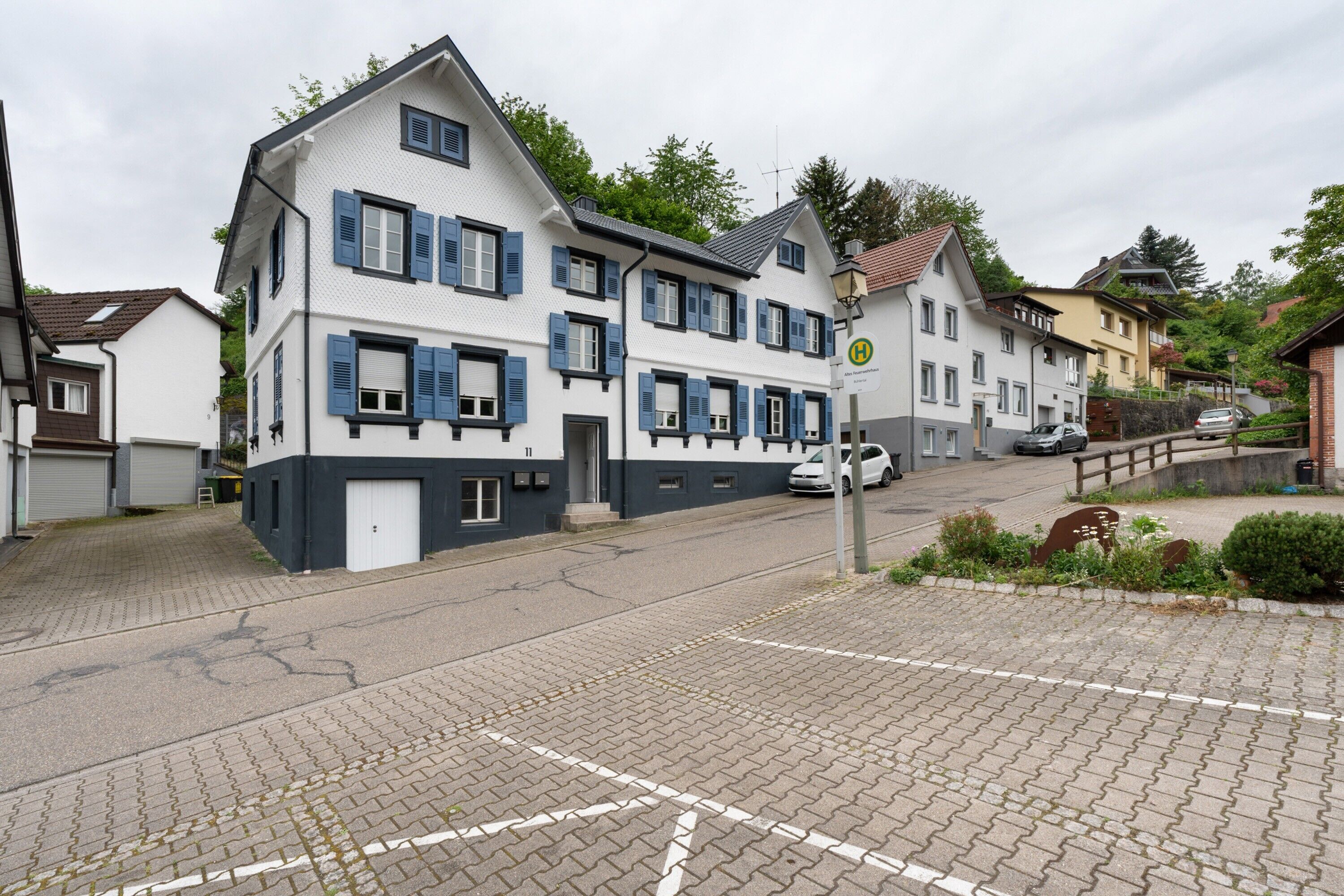 Un immeuble à plusieurs étages, doté d’une façade blanche et de volets bleus, une rue pavée et une voiture garée.