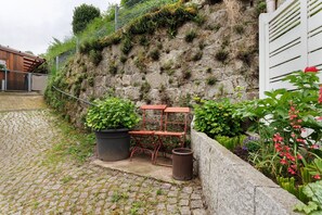 A cobblestone pathway leading to a rustic outdoor seating area with a metal table and chairs, surrounded by lush greenery and potted plants.