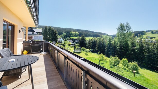 A balcony with a table and chairs, a view of a green landscape, and a building in the background.