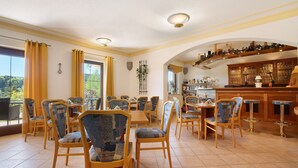 A dining area with wooden chairs and tables, a bar with stools, and a view of greenery outside.
