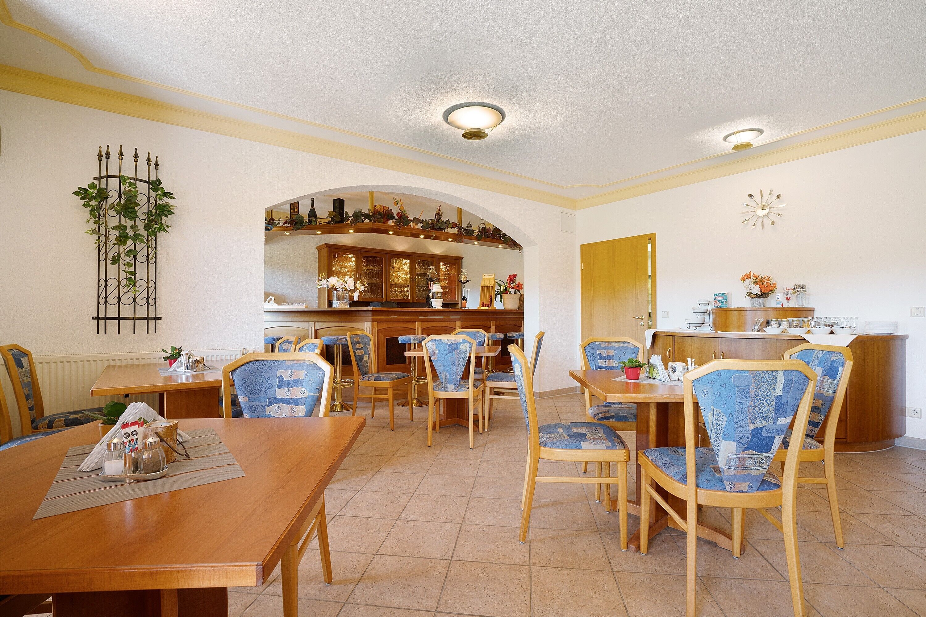 Un coin repas avec des tables et des chaises en bois, une cuisine à l’arrière-plan et une plante décorative accrochée au mur.