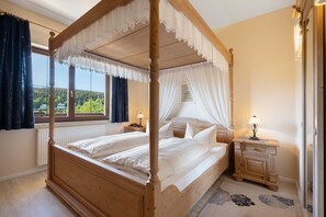 A four-poster bed with white bedding, a wooden nightstand with a lamp, and a view of greenery outside.
