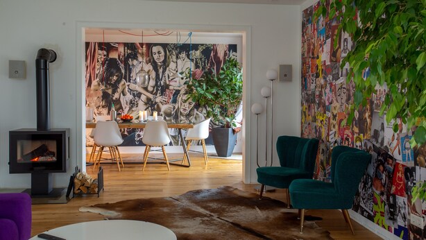 Een moderne woonkamer met een open haard, een eethoek met stoelen, een bank, een groene fauteuil en een muur bedekt met een fotocollage.