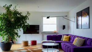 Een moderne woonkamer met een paarse bank, een grote potplant, een ronde salontafel en een flatscreen tv die aan de muur is bevestigd.