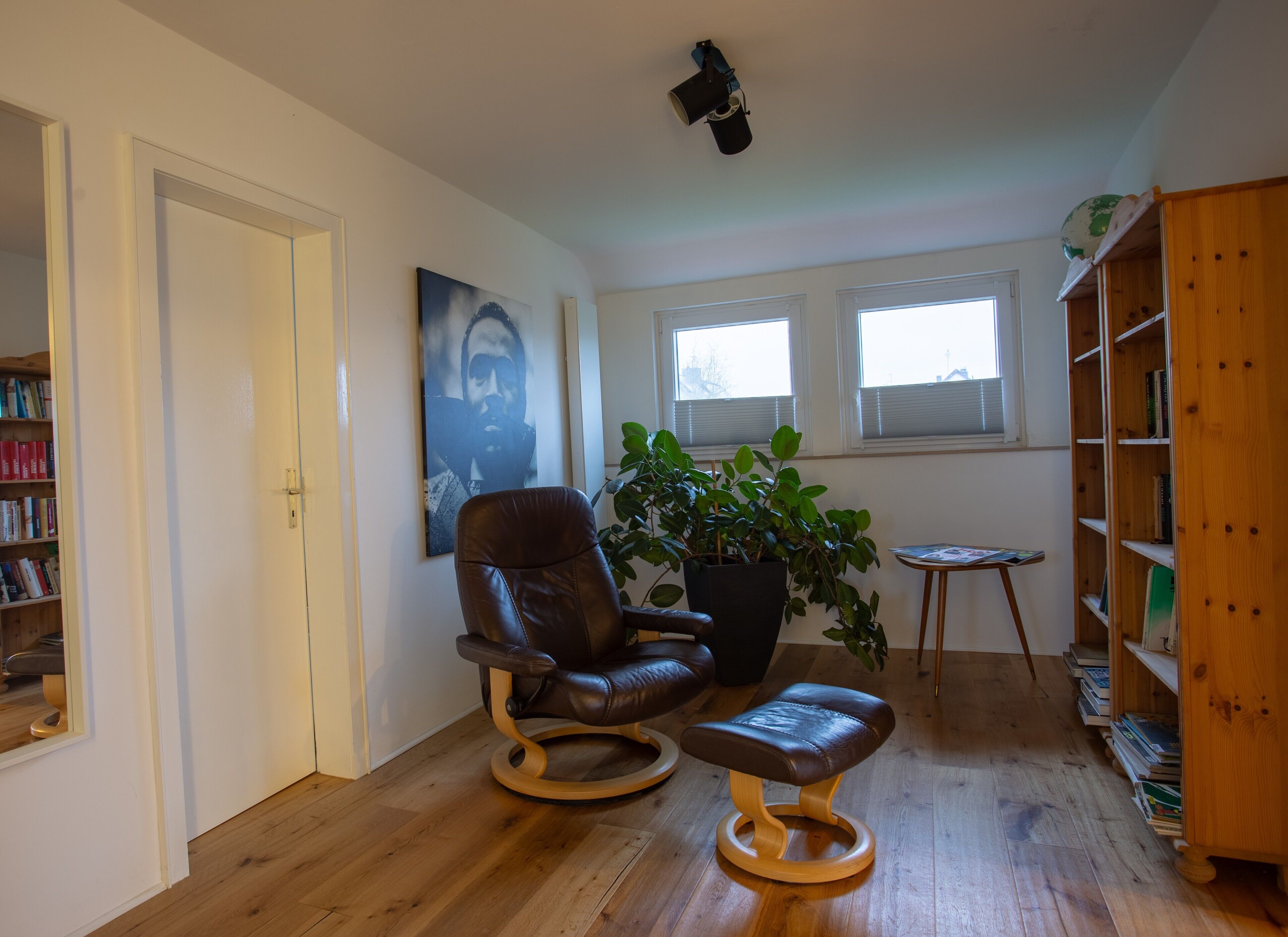 Une pièce comprenant un fauteuil en cuir, une table d’appoint en bois, une bibliothèque, une plante en pot et un tableau accroché au mur.