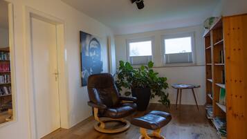 Une pièce comprenant un fauteuil en cuir, une table d’appoint en bois, une bibliothèque, une plante en pot et un tableau accroché au mur.