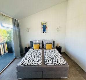 A bedroom with a bed, two bedside tables, a painting, and a view of the outdoors.