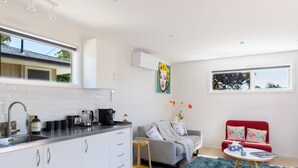 A modern kitchen with white cabinets, a stainless steel sink, and a coffee maker. There is a grey sofa with colorful cushions, a red armchair, and a small round table with a blue rug. - Classy Modern Unit.  Walk to shops, bars & cafes.  Pet-Friendly! Bus at gate. (Christchurch)