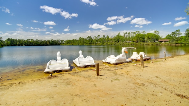 A sandy beach with several white paddle boats docked near the water's edge, surrounded by a forested area and a clear blue sky. - Biloxi Resort Cottage w/ Pool & Lake Access! (Biloxi)