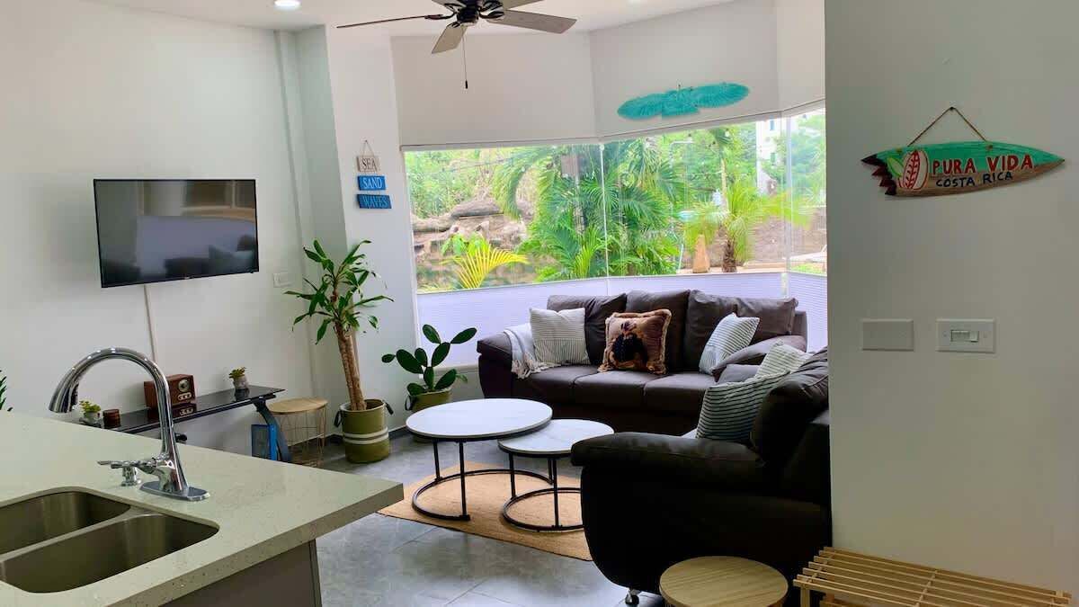 A modern living room with a sofa, coffee table, and TV. There's a kitchen area with a sink and a dining table. The room has large windows with a view of greenery outside.