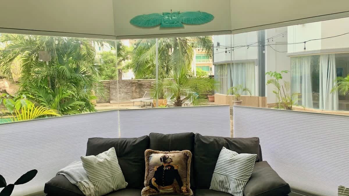 Une pièce avec un canapé en cuir noir, un coussin orné d’un motif de chien et une vue sur un jardin avec des palmiers et un mur en pierre.