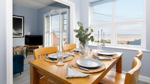 A dining area with a wooden table set for four, a television in the background, and a view of the beach through the window. - Chic 2-Bed Seafront Haven, Near Felpham Beach (Felpham)