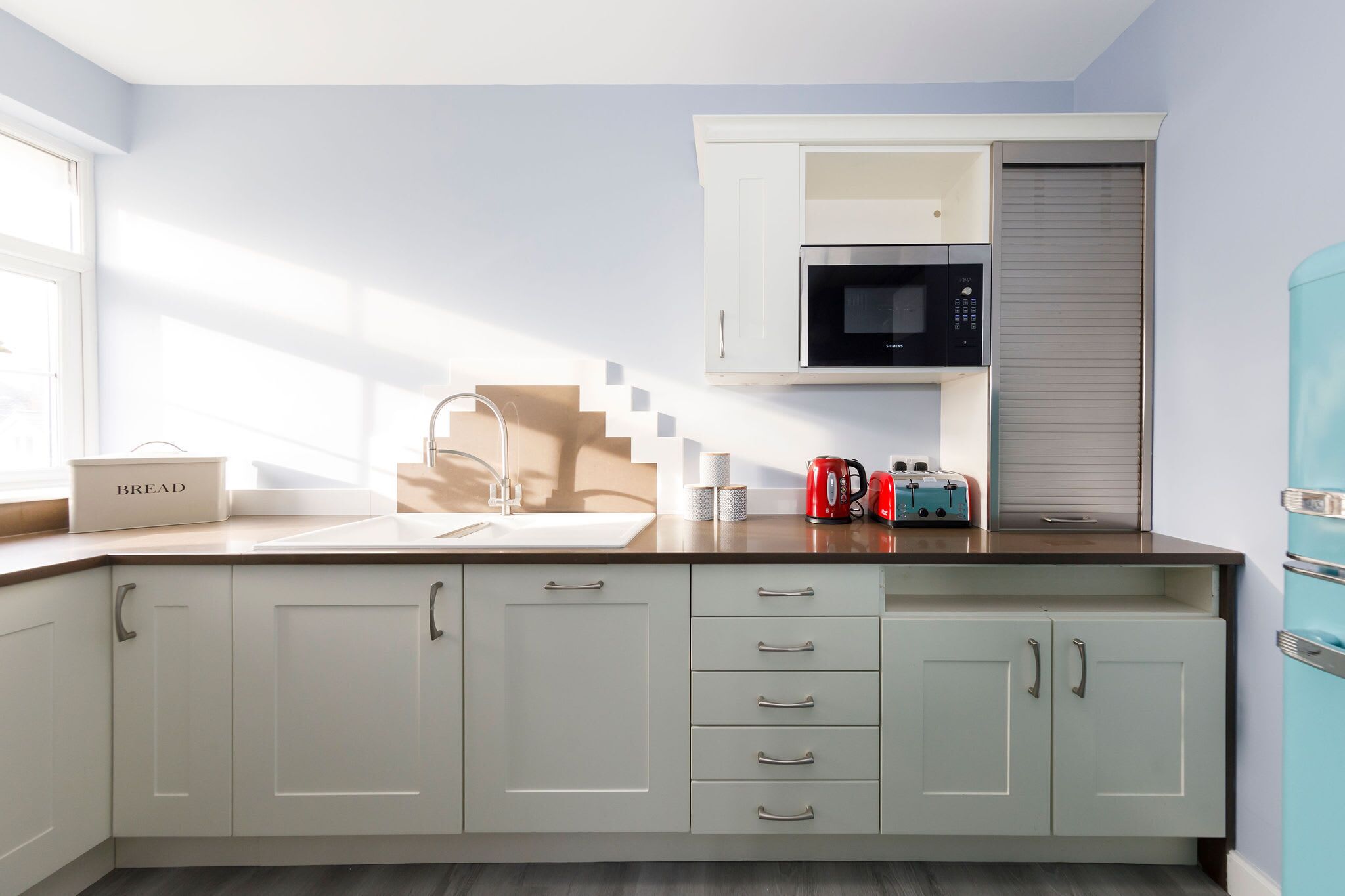 Una cocina con armarios blancos, microondas, hervidor y panera.