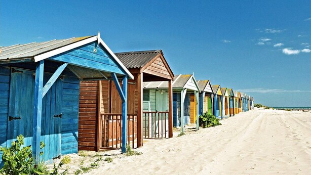 A row of beach huts with sloped roofs, wooden doors, and a sandy beach in the background. - Chic 2-Bed Seafront Haven, Near Felpham Beach (Felpham)