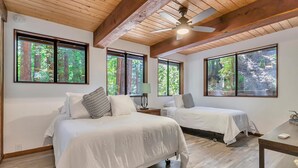 Una habitación con dos camas, ventanas amplias con vistas al bosque, vigas de madera en el techo y un ventilador de techo.