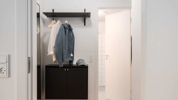 A hallway with a black wardrobe, a coat rack with clothes, and a door leading to a bathroom.