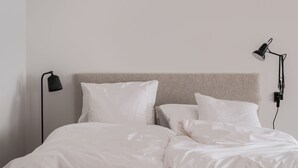 A neatly made bed with white bedding, a headboard, and a black floor lamp on the left.