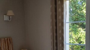 Een slaapkamer met een bed, een nachtkastje met lamp, een raam met gordijnen en uitzicht op groen buiten.