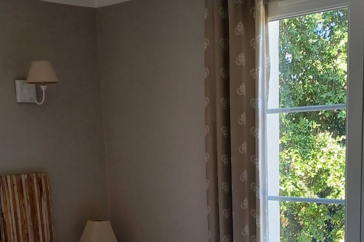 A bedroom with a bed, a nightstand with a lamp, a window with curtains, and a view of greenery outside.