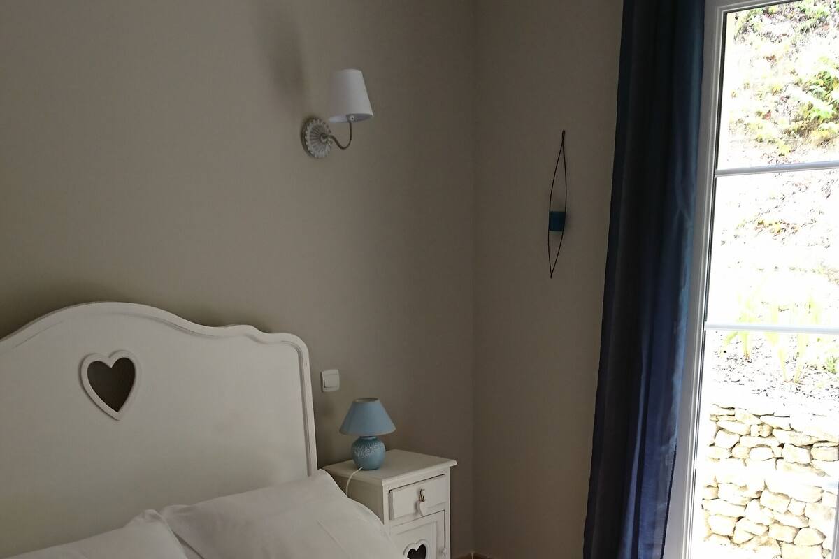 A bedroom with a bed, a nightstand with a lamp, a wall-mounted mirror, and a window with curtains.