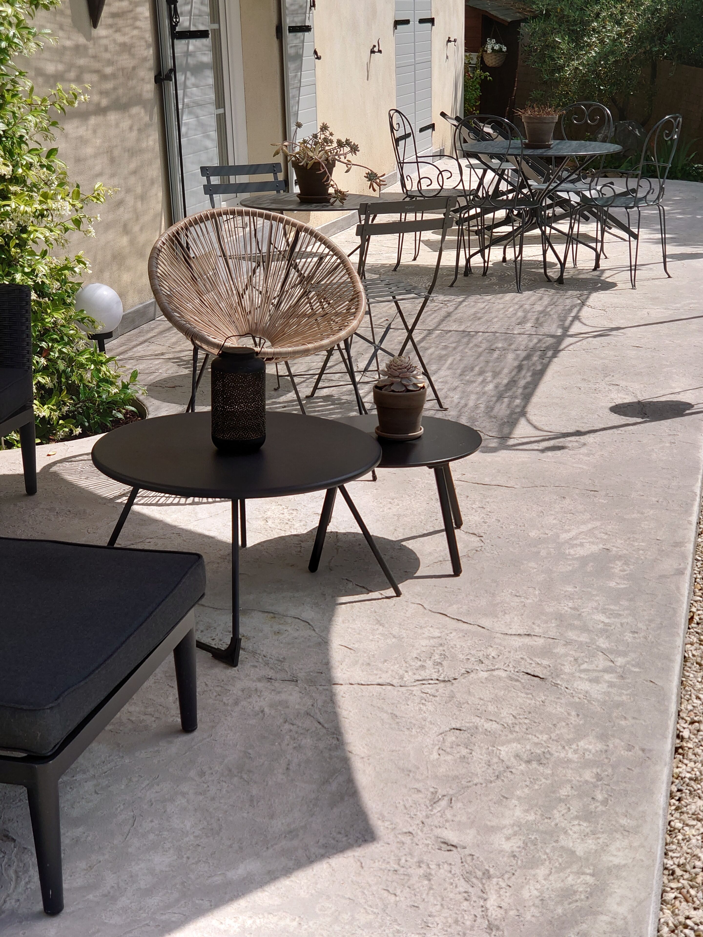 An outdoor patio area with round tables and wicker chairs, potted plants, and a concrete floor.