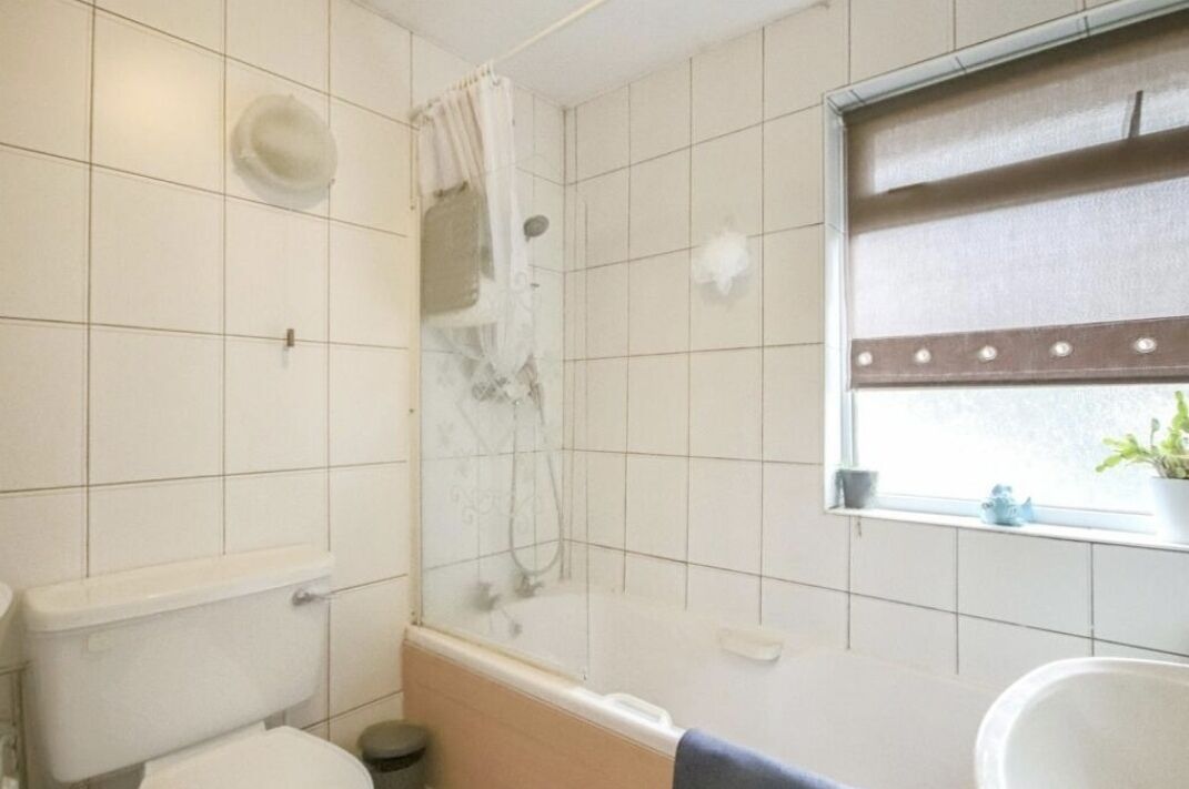 Un baño con paredes de azulejos blancos, zona de ducha, inodoro y ventana con persianas.
