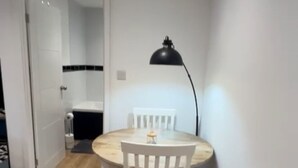 A small, well-lit room with a round wooden table and two white chairs. A black floor lamp is on the table, and a white radiator is on the wall.