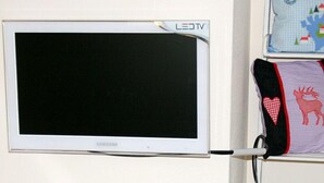 Un televisor blanco de Samsung montado en una pared, con un cojín azul que muestra un diseño de ciervo y corazón sobre una estantería.