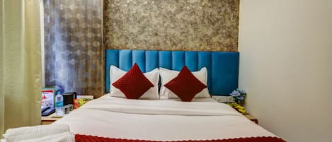 Une chambre d’hôtel avec un lit, une couvre-lit rouge matelassé, deux oreillers rouges, une couverture blanche, une tête de lit bleue, un mur avec un motif texturé, une table de chevet avec un téléphone, une bouteille d’eau, un menu et un arrangement floral.
