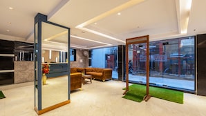 A modern hotel lobby with a sofa, a mirror, and a view of the street through large windows. - Hotel Stay House Inn (New Delhi)