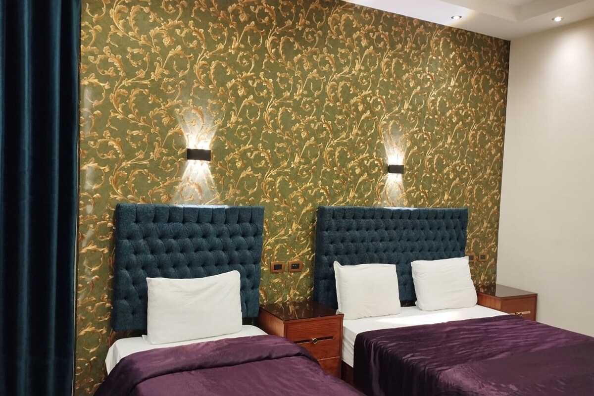 Habitación de hotel con dos camas, cabeceros azul oscuro, almohadas blancas, colchas púrpuras y un papel tapiz verde estampado con detalles dorados.