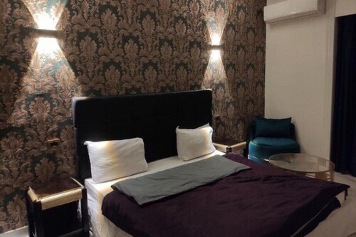 A hotel room with a large bed, two bedside tables, a round glass table, a dark headboard, patterned wallpaper, and two wall-mounted lights.