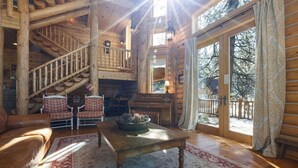 A log cabin interior with a fireplace, a wooden staircase, and a large window overlooking a snowy landscape.