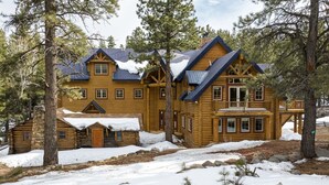 A log cabin with a balcony and a stone extension, surrounded by snow and trees.