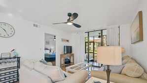 A living room with a ceiling fan, a clock, a sofa, a coffee table, and a TV. - Bradenton Beach Condo -2 Pools- 15 minutes to Anna Maria Island Beaches-First Floor- End Unit (Bradenton)