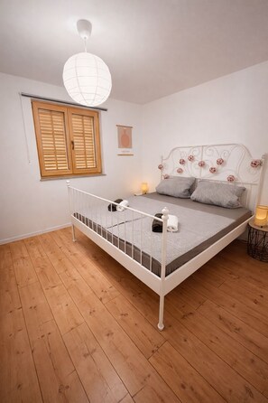 A bedroom with a white metal bed frame, a wooden floor, a white ceiling light, a window with wooden shutters, and a wall-mounted picture.