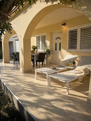 An outdoor patio with a table and chairs, a lounge chair, and a door with shutters.