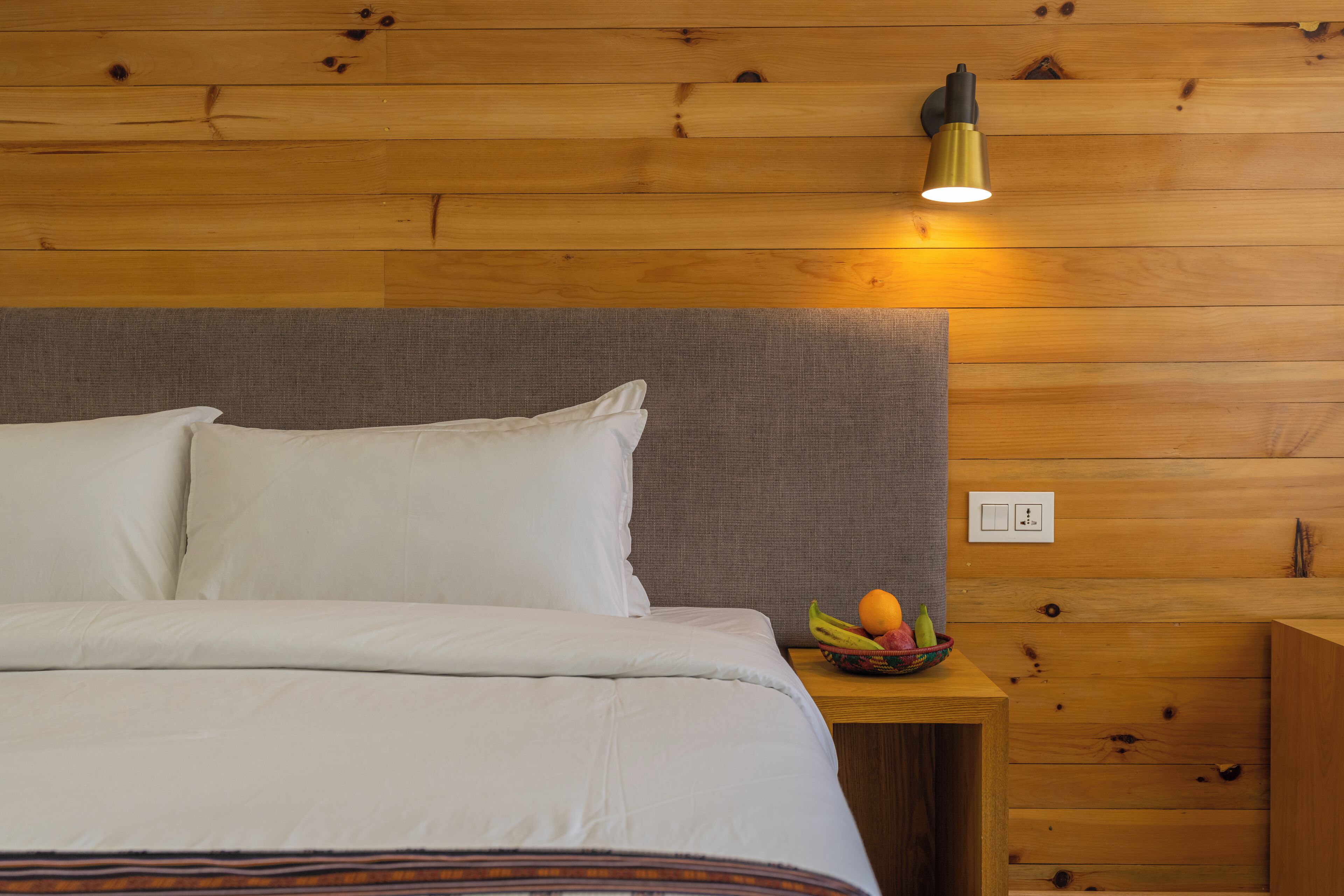Un lit bien fait, avec du linge de lit blanc, une tête de lit grise et une petite table de chevet en bois sur laquelle se trouve un panier de fruits.