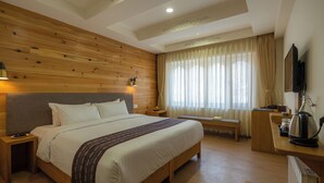A hotel room with a wooden headboard, a large bed, a bedside table with a lamp, a bench, a desk with a kettle, and a TV mounted on the wall. - The Willows (Thimphu)