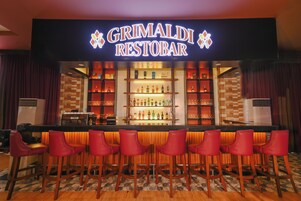 Zona de bar con un letrero que dice "GRIMALDI RESTOBAR", que incluye una barra, varias estanterías con botellas y seis taburetes rojos.