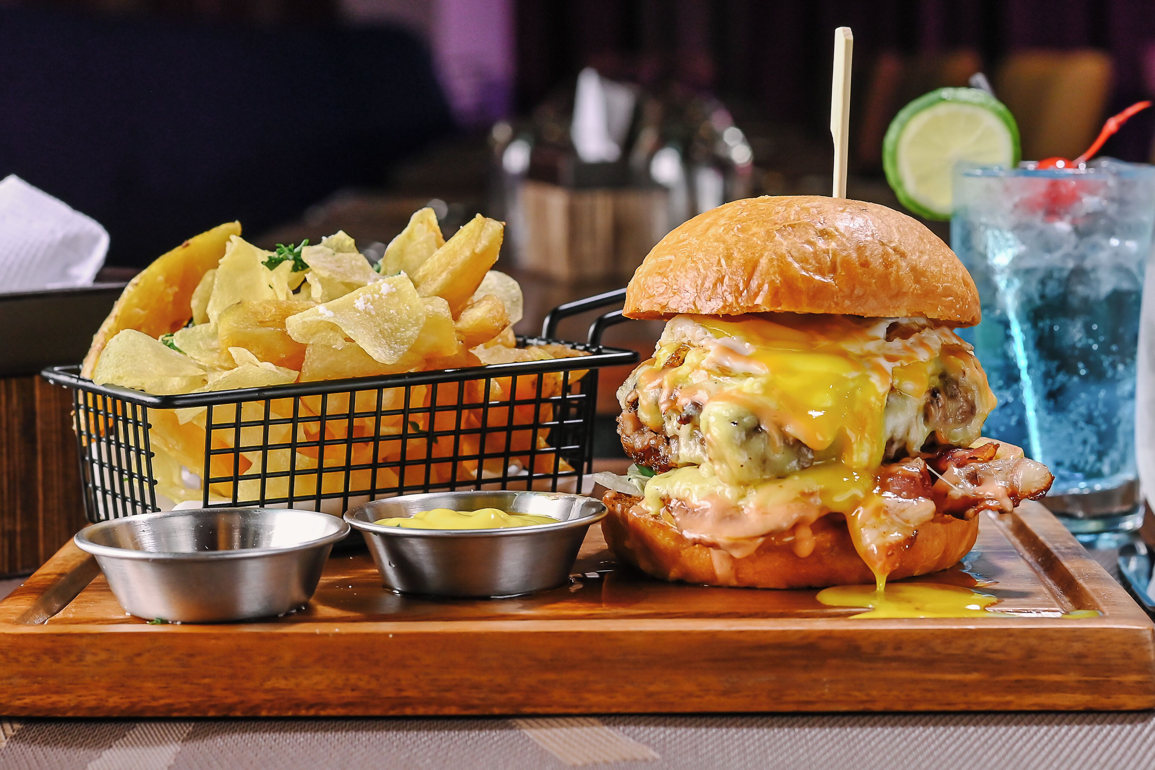A burger with cheese and bacon, a basket of chips, and two small containers of condiments on a wooden tray.