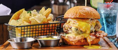 A burger with cheese and bacon, a basket of chips, and two small containers of condiments on a wooden tray.