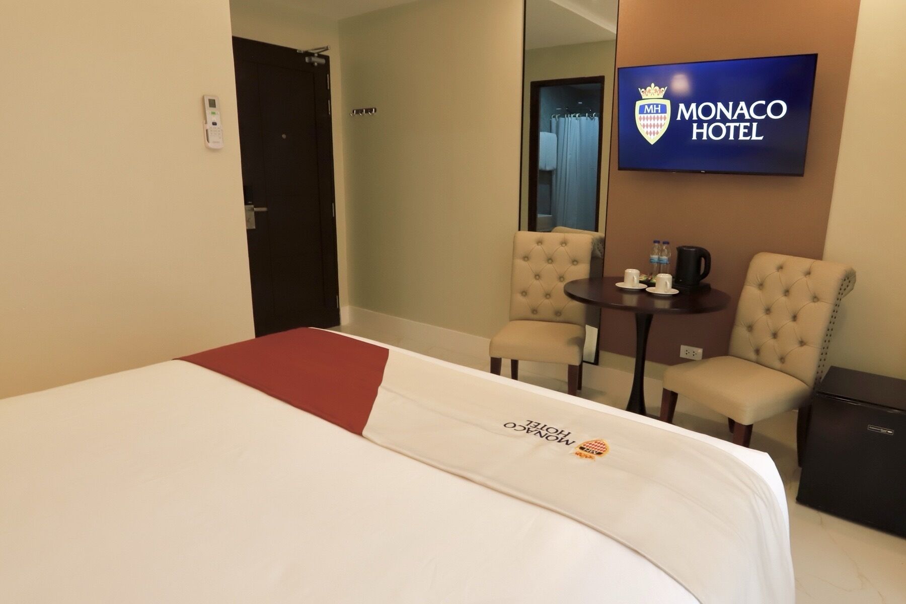 A hotel room with a bed, two armchairs, a small table with a kettle, and a television displaying the hotel name.